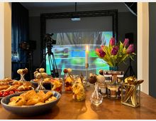 Ein Tisch mit verschiedenen Snacks und Desserts, darunter Croissants, Obst in Gläsern und Cake-Pops, steht im Vordergrund. Im Hintergrund sind Filmkameras, eine Leinwand mit einer Projektion und ein Kerzenleuchter sowie ein Strauß Tulpen sichtbar.