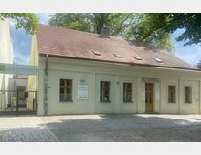 Ein einstöckiges Gebäude mit beigem Putz und roten Ziegeldach, das mehrere Fenster mit Holzrahmen und ein Holzeingangstür besitzt, gekennzeichnet als „POTTEN Salon Beauté“. Links daneben befindet sich ein Eingangstor mit einem Schild für „fitbox – Die Fitness Revolution“.