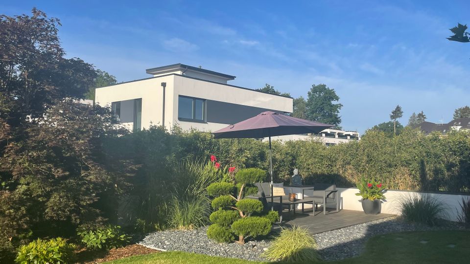 Garten mit Rasen und bepflanztem Kiesbeet, daneben eine kleine Holzterrasse mit Sitzgruppe und Sonnenschirm, umgeben von Hecken und Ziersträuchern. Im Hintergrund steht ein modernes, weißes Flachdachhaus mit großen Fenstern vor blauem Himmel.