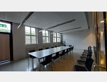 Ein Konferenzraum mit einem langen rechteckigen Tisch, umgeben von schwarzen Stühlen. Der Raum hat große Fenster, eine weiße Decke mit länglichen Hängelampen und helle Wände; im Hintergrund steht eine Zimmerpflanze.