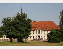 Herrenhaus mit heller Fassade und rotem Satteldach, umgeben von grünen Bäumen und einer Wiese im Vordergrund, bei klarem Himmel.