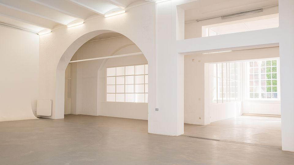 Heller, leerer Innenraum mit weißen Wänden, hohen Decken und einem großen Rundbogen, der zwei Bereiche verbindet. Große Sprossenfenster lassen viel Tageslicht auf den grauen Betonboden; an der Decke sind lineare Leuchten montiert.