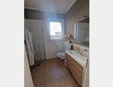 Ein modernes Badezimmer mit dunklem Holzboden, bodentiefer Glasdusche links, einem wandhängenden WC unter einem Fenster und einem Waschbecken mit Holzunterschrank rechts. Die Wände sind in Weiß und Grau gestaltet.