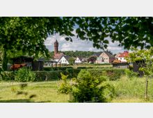 Eine ländliche Szene mit gepflegten Gärten im Vordergrund, kleinen Gartenhäusern und einer Reihe von Wohnhäusern im Hintergrund. Ein roter Backsteinturm mit spitzer Spitze überragt die Landschaft, umgeben von Bäumen und Wiesen.