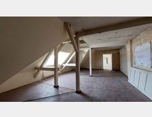 Ein unausgebauter Dachbodenraum mit sichtbaren Holzstützen und schrägen Wänden. Der Boden ist roh, die Wände sind teilweise verputzt, und links befinden sich Dachfenster, die natürliches Licht hereinlassen.