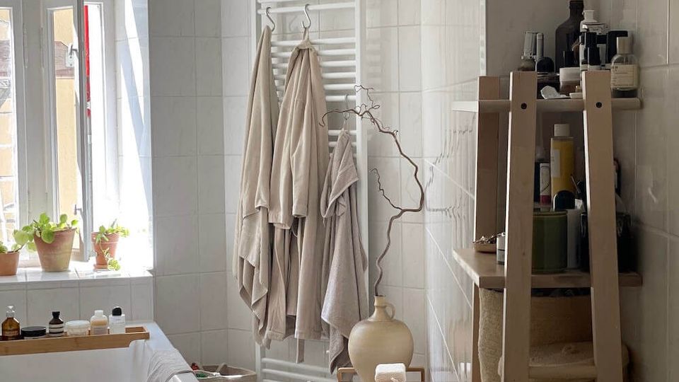 Bright white-tiled bathroom with a bathtub under a tall window, small potted plants on the sill, and a towel radiator holding hanging robes. A wooden ladder-style shelf with toiletries and baskets stands beside a small stool and decorative vase on the tiled floor.