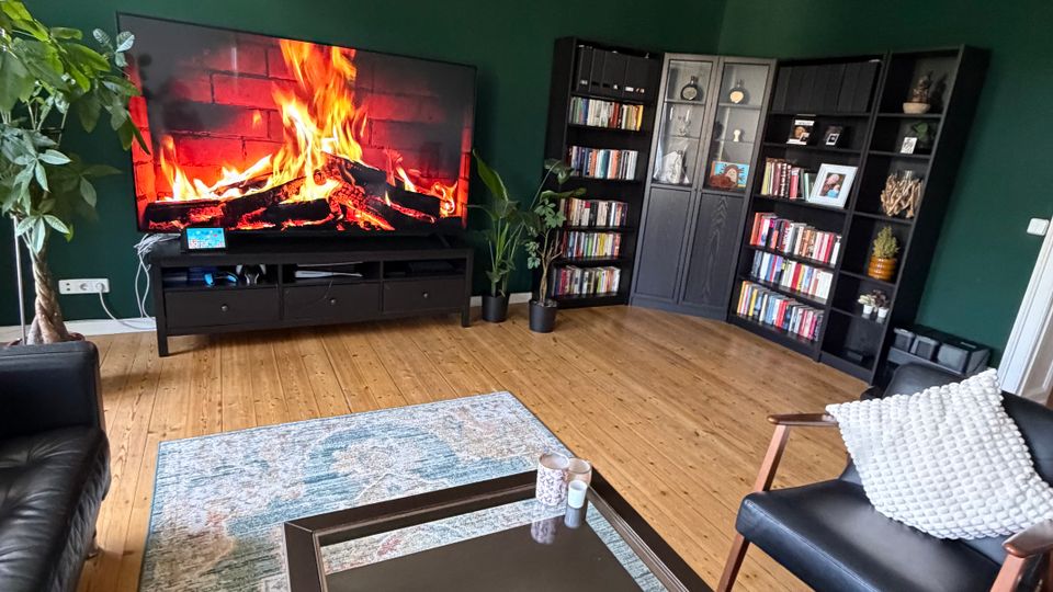 Wohnzimmer mit dunkelgrünen Wänden und Holzdielenboden, mit großem Fernseher auf niedrigem Sideboard, der ein Kaminfeuer zeigt. In der Ecke stehen schwarze Bücherregale mit Schrankelement, davor ein Teppich und ein Glastisch; darüber hängt eine Deckenleuchte mit mehreren Glaszylindern.