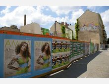 Eine Straße in einer städtischen Umgebung mit Bauzäunen, die mit verschiedenen Plakaten und Werbung bedeckt sind. Im Hintergrund befinden sich mehrstöckige Gebäude mit teils sichtbaren Graffitis sowie ein Schornstein, unter einem teils bewölkten Himmel.