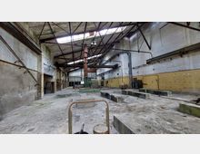 Abbildung einer verlassenen Industriehalle mit hohen Decken und sichtbaren Stahlträgern. Der Raum ist heruntergekommen, zeigt bröckelnde Wände und vereinzelt moosbewachsene Stellen; Teile der Wand sind gelb gestrichen, und die Halle enthält verschiedene veraltete Strukturen und Maschinenreste.