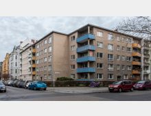 Mehrstöckiges Wohnhaus in einer urbanen Umgebung, mit einer braunen Fassade und Balkonen in Blau und Gelb. Vor dem Gebäude befinden sich geparkte Autos entlang einer Wohnstraße, sowie Gehwege und ein Straßenschild.