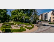 Kleiner städtischer Park mit symmetrischen, von Hecken gesäumten Wegen, einer kreisförmigen Sitzanlage in der Mitte und umgeben von hohen Bäumen. Im Hintergrund befinden sich Wohngebäude und rechts führt eine halbrunde Treppe zu einer gepflasterten Fläche.