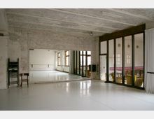 Industrie-inspirierter Tanz- oder Bewegungssaal mit weißen Wänden, Spiegeln an einer Wand und großen Fenstern mit Holzrahmen. Der Boden besteht aus glänzendem weißem Material, und eine Trennwand aus Glas mit Vorhängen trennt den Raum von einem angrenzenden Bereich.