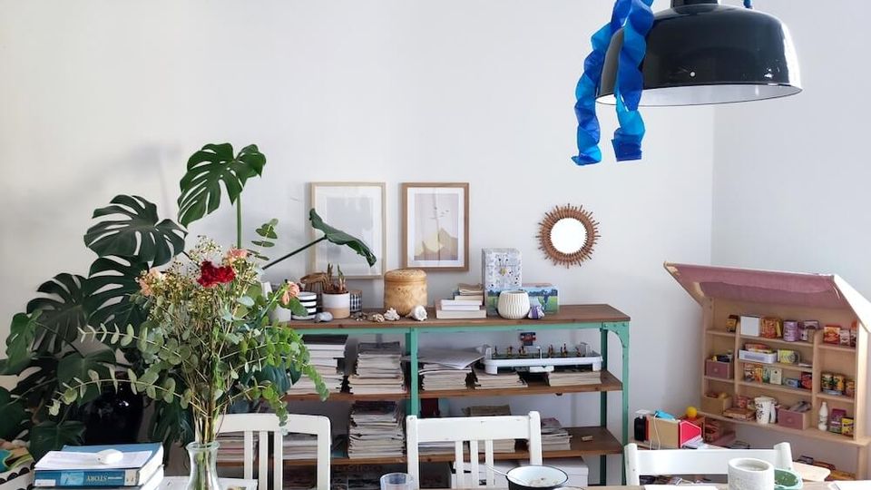 A casual dining or workspace with a wooden table surrounded by mismatched chairs, cluttered with books, papers, and craft items. The background features a white wall, a green shelving unit filled with books and decor, a large leafy plant, and a hanging black lamp with blue streamers.