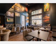Innenraum eines stilvollen, modern eingerichteten Cafés mit dunkelblauen Wänden, dekoriert mit zahlreichen Kunstwerken und Fotodrucken. Die Bestuhlung besteht aus gepolsterten Sesseln und Stühlen an dunklen Tischen; durch ein großes Fenster fällt Tageslicht herein und bietet einen Blick auf ein historisches Gebäude mit Säulen.