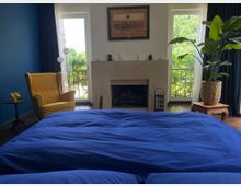 Schlafzimmer mit großem Bett in dunkelblauer Bettwäsche im Vordergrund und einem Kamin mit hellem Steinmantel an der Stirnwand. Links steht ein gelber Ohrensessel vor einer blauen Wand, rechts eine große Zimmerpflanze; zwei hohe Fenstertüren mit Vorhängen öffnen den Blick ins Grüne.