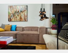 Wohnzimmer mit einer großen, beigefarbenen Couch, farbigen Kissen und einer modernen Pendelleuchte. An der Wand hängt ein buntes abstraktes Gemälde, daneben steht eine Pflanze in einer roten Vase auf weißen Beistelltischen; im Vordergrund ein Couchtisch mit Büchern.