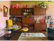Wohnzimmer mit pastellrosa Wänden und einem grünen Deckenrand. Ein gelber Sessel mit passendem Hocker steht vor einem vintage Holzregal mit Büchern, Dekorationsgegenständen und Schubladen. Auf dem Boden liegt ein bunter Teppich, daneben ein kleiner Tisch mit einer Vase.