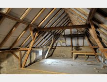 Holzrahmen-Dachboden mit sichtbarer Holzkonstruktion und einem kleinen Fenster an der Seitenwand. Ein Kronleuchter hängt von der Decke, und der Raum ist größtenteils leer, mit einem rohen Fußboden und unverputzten Wänden.