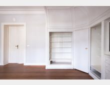 Heller, leerer Innenraum mit weiß gestrichenen Wänden, hoher Decke mit Stuck und dunklem Holzboden. Zu sehen sind eine Tür sowie ein eingebauter Wandschrank mit offenen Regalböden und geöffneter Schranktür.