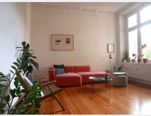 Wohnzimmer mit Holzboden, rosa Sofa, rundem Couchtisch und modernen Möbeln. Große Fenster lassen Tageslicht herein, auf der Fensterbank stehen verschiedene Pflanzen in Töpfen.