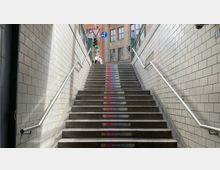 Treppenaufgang einer U-Bahn-Station mit hellen Fliesenwänden und Edelstahlhandläufen. Auf den Stufen sind bunte Textabschnitte zu sehen, die stilisierte Botschaften tragen, und im Hintergrund ist ein Straßenausgang mit Ampeln und Verkehrszeichen sichtbar.