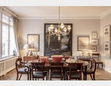 Esszimmer mit klassischem Stil, das einen dunklen Holztisch und Stühle zeigt. An der Wand hängen ein großes Landschaftsgemälde und mehrere kleinere Kunstwerke; ein Kronleuchter beleuchtet den Raum, der an einer Fensterfront mit Vorhängen gelegen ist.