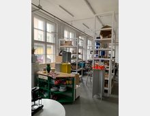 Heller Werkraum mit großen Fenstern, ausgestattet mit Regalen, die mit verschiedenen Utensilien und Materialien gefüllt sind. Im Vordergrund befinden sich eine grüne Küchenablage mit Geschirr sowie eine Arbeitsplatte, im Hintergrund weitere Arbeitsbereiche.