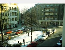 Straßenszene in einem Wohnviertel mit mehrstöckigen Wohngebäuden im klassischen Stil, umliegenden Bäumen und schneebedeckten Grünflächen. Geparkte Autos säumen die Straße, Passanten und ein Fotograf befinden sich im Vordergrund.