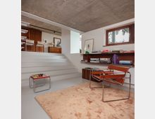 Modern split-level living room with wide white steps leading to a raised seating area, exposed concrete ceiling, and built-in wood shelving filled with books beneath a small window. Minimalist furniture includes chrome-framed chairs and a low table on a large beige rug.