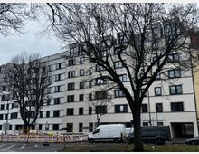 Straßenszene mit einem langen, mehrstöckigen Wohn- oder Bürogebäude mit hellen Fassaden und vielen gleichmäßigen Fenstern, teilweise mit Dachgauben. Im Vordergrund stehen kahle Bäume sowie Baustellenabsperrungen und Transporter entlang der Straße.