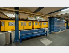 U-Bahnhof mit einer gelb lackierten U-Bahn der Linie U1 an der Station "Uhlandstraße". Im Vordergrund stehen eine blaue Sitzbank, eine Mülltonne und eine blau geflieste Säule, umgeben von gewölbten Decken und Beleuchtung.