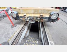 Ein niedriger Wartungsgraben in einer Werkstatt mit Schienen und einer Arbeitsplattform darüber. Im Vordergrund sind gelb-schwarz markierte Stufen, und im Hintergrund ist das Ende eines Fahrzeugs mit großen Scheinwerfern sichtbar.