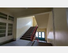 Treppenhaus mit einfachen, gelb-beigen Wänden, einer Treppe mit dunklen Stufen und einem roten Geländer. Im Hintergrund befinden sich eine Glastür mit grünem Rahmen und ein Notausgangsschild darüber.