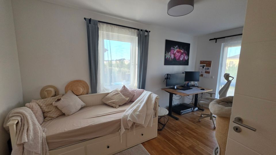 Helles Schlafzimmer mit Tagesbett, Kissen und Decken neben einem Fenster mit grauen Vorhängen auf Holzfußboden. Gegenüber steht ein Arbeitsplatz mit Schreibtisch, Monitoren und Bürostuhl; ein weiteres Fenster und ein Bild hängen an der Wand.