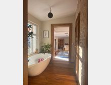 Ein helles Badezimmer mit Holzfußboden, einer freistehenden weißen Badewanne und Pflanzen. Im Hintergrund ist durch einen Türrahmen ein weiterer Raum mit Schränken und einer hellen Matte erkennbar.