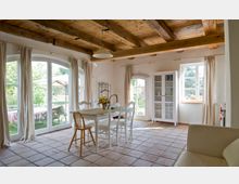 Esszimmer in einem Landhausstil mit weißem Holztisch, passenden Stühlen und einem Holzhocker. Große Fenster und Glastüren lassen Blick auf einen Garten zu, Zimmerdecke aus sichtbaren Holzbalken und Fliesenboden mit Terrakotta-Optik.