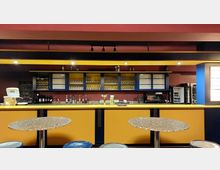 Eine Bar mit gelb-blauer Einrichtung vor einer roten Wand. Im Hintergrund befinden sich Regale mit Gläsern, Flaschen und Utensilien, während im Vordergrund runde Tische und Hocker zu sehen sind.