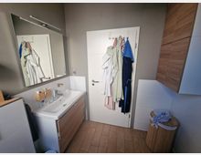 Modernes Badezimmer mit einer weißen Tür, an der mehrere Bademäntel hängen. Links ein Waschbecken mit Holzunterschrank und großem Wandspiegel, rechts ein Holzschrank und ein Wäschekorb. Der Boden besteht aus hellbraunen Fliesen.