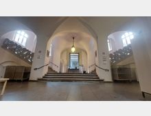 Eingangsbereich eines historischen Gebäudes mit einer zentralen Treppe, flankiert von zwei geschwungenen Seitentreppen mit verzierten Geländern. Hohe Rundbögen, große Fenster und eine zentrale Hängelampe prägen die helle Architektur.