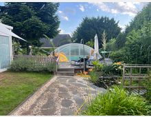 Garten mit gepflastertem Weg, kleinen Kiesflächen und bepflanzten Beeten. Im Hintergrund ein Pool mit gewölbter Überdachung, umgeben von Terrasse, Gartenmöbeln und großen Büschen.