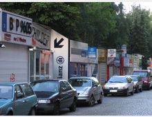 Straße mit geparkten Autos vor einer Reihe kleiner Geschäfte, die hauptsächlich Schilderherstellung und Dienstleistungen anbieten; die Läden haben farbige Werbeschilder mit Text, während Bäume den Hintergrund bilden.