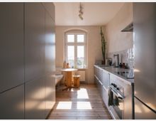 Moderne, helle Küche mit Holzfußboden, einem großen Fenster im Hintergrund und einer schlichten Einrichtung. Auf der rechten Seite befindet sich eine Arbeitsfläche mit Spüle und Herd, links sind glatte, graue Küchenschränke zu sehen, und im hinteren Bereich steht ein runder Holztisch mit Sitzbank.