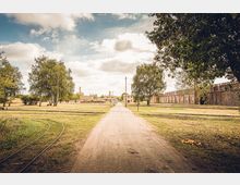 Weitläufiges, parkähnliches Gelände mit sandigem Weg in der Mitte, umgeben von Bäumen und Wiesen; im Hintergrund sind industrielle Backsteingebäude, Schornsteine und alte Schienenstränge zu sehen.