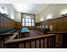 Gerichtssaal mit Holzwandverkleidung und verzierten Deckenstuck. Der Raum enthält eine Richterbank, zwei Tische mit grünen Tischauflagen und Sitzreihen mit gepolsterten Stühlen, sowie ein großes Fenster im Hintergrund.