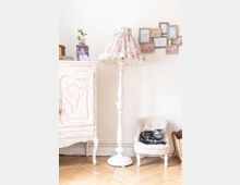 Ein hell eingerichtetes Wohnzimmer mit einem cremefarbenen, antik wirkenden Schrank, einer Stehlampe mit geblümtem Lampenschirm und einem kleinen Polstersessel, auf dem eine getigerte Katze liegt. Im Hintergrund hängen mehrere kleine, dekorative Bilderrahmen an der weißen Wand.