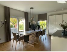 Esszimmer in einem modernen Wohnhaus mit einem großen Holztisch und sechs Stühlen. Große Fenster mit Vorhängen und Blick auf einen Garten sorgen für viel Tageslicht; links steht ein Metall-Kerzenständer, rechts eine schlichte weiße Küchenfront.