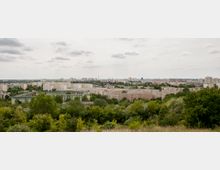 Blick auf eine städtische Wohnsiedlung mit flachen, rechteckigen Wohnblöcken, umgeben von Bäumen und Grünflächen, im Hintergrund weitere Hochhäuser und eine weite, urbane Landschaft unter bewölktem Himmel.