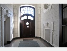 Eingangsbereich mit einer dunklen Holztür und rundem Fenster in der Mitte, umgeben von weißen Wänden mit dekorativen Stuckelementen. Die Tür hat ein oben halbkreisförmiges Glasfenster, und der Boden ist mit einem Teppichläufer ausgelegt.