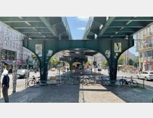 Eine belebte Straßenkreuzung in der Stadt mit einer grünen, metallischen Hochbahnbrücke, unter der Fahrräder abgestellt sind. Im Hintergrund sind Wohnhäuser, Ladenfassaden, Verkehrsschilder und eine belebte Fußgängerzone zu sehen.