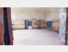 Verlassener Raum mit abblätterndem Putz an den Wänden, auf denen bunte Graffiti sichtbar sind. Der Boden ist staubig, und eine offene Tür führt zu einem weiteren Raum, der ebenfalls Graffiti zeigt.